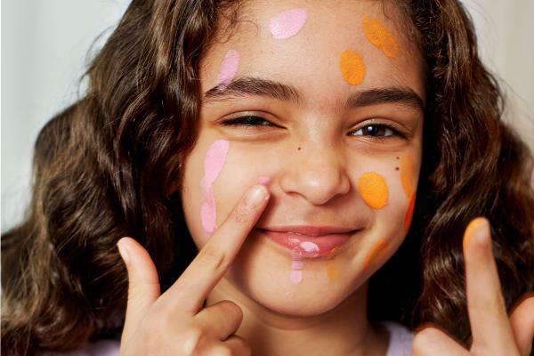 Young brown haired girl with a playful expression finger painting her face pink and orange
