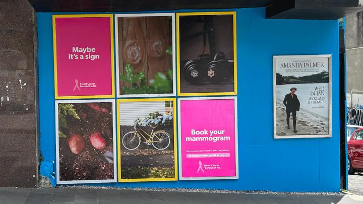 A series of outdoor advertisements against a blue construction wall. 4 pictures depict: two circular patterns in plans of wood; a bicycle; two potatoes on the ground; and two dumbbells. One poster says "Maybe it's a Sign", and one poster says "Book your Mammogram" 