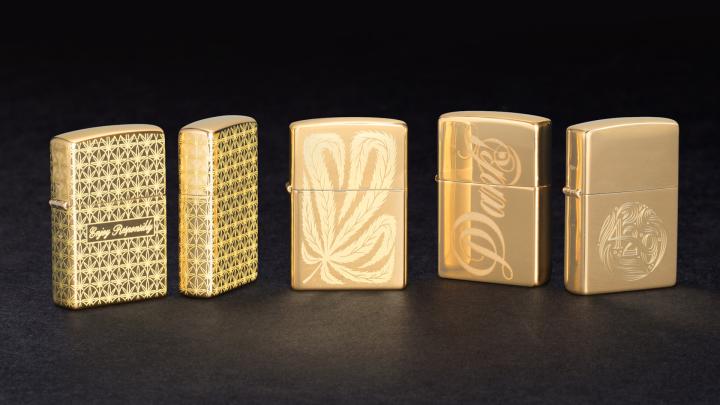 5 gold Zippo lighters standing upright against a black background. This is the High-class Collection. the first two feature an engraved marijuana leaf pattern, with "enjoy responsibly" engraved in script. The middle one has one large marijuana leaf engraved into it. the 4th has the word "Dank" engraved in script across the face. The far right one has "420" engraved on it in a circular, continuous font and pattern