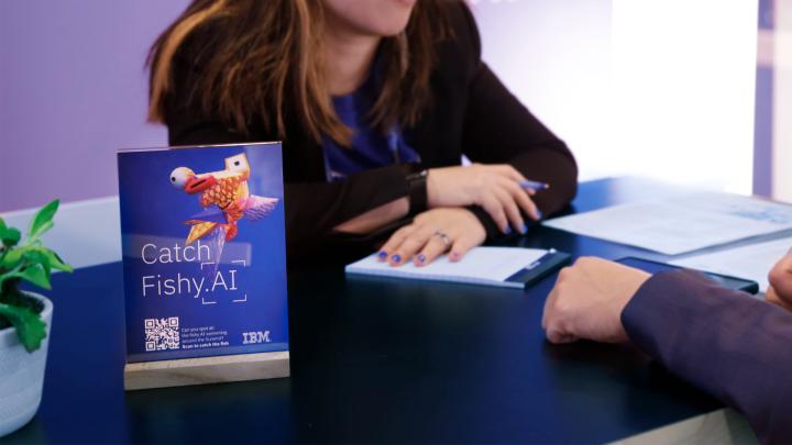 Woman sitting at a desk at the IBM activation booth at Adobe Summit, on the table is a display featuring AI generated image of a messed up fish with the label "Catch Fishy.AI" 