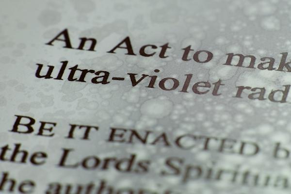 A close up of text from the UK's Sunbeds Regulation Act. We can see the words "An act to" and "ultra-violet" and "Be it enacted". The paper has been put through the radiation of a sunbed so it is marked with burns and discolorations