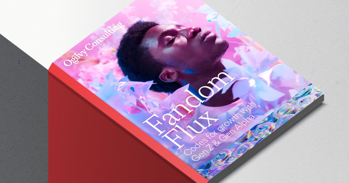 A report cover from Ogilvy Consulting, titled 'Fandom Flux' with the subtitle 'Codes for growth with Gen Z & Gen Alpha.' The cover features a person reclining with closed eyes, surrounded by shimmering, iridescent elements in shades of pink, purple, and blue, suggesting a dreamy or futuristic aesthetic. The report is angled on a grey and white surface, showing its red spine.