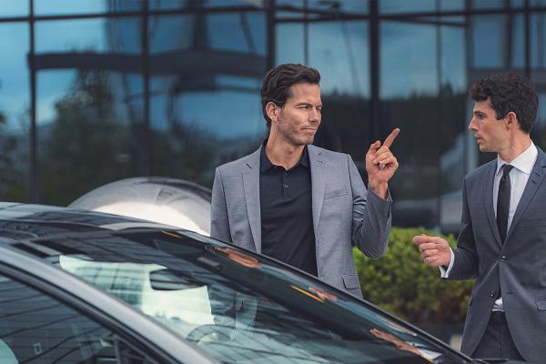 Two young men standing behind an Audi car. They are both wearing suits. The man on the left is point his finger at the man on the right.