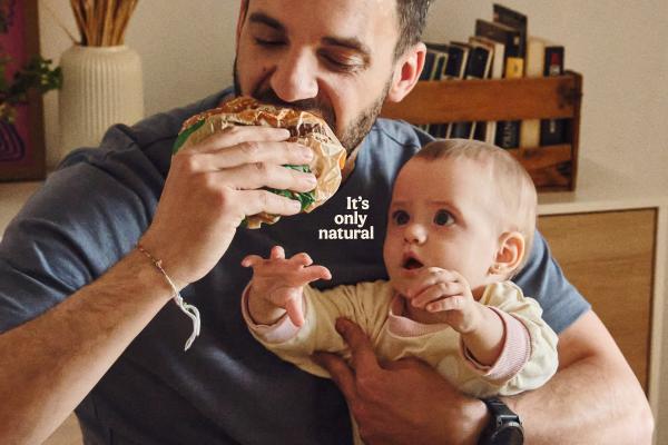 It's Only Natural — Burger King A man with a beard is holding a baby and taking a large bite out of a burger wrapped in paper. The baby, held in his left arm, looks up at the burger with wide, curious eyes and has its hands reaching out. White text on the image reads "It's only natural". The background shows a warm, indoor setting with a vase and a wooden crate filled with books.