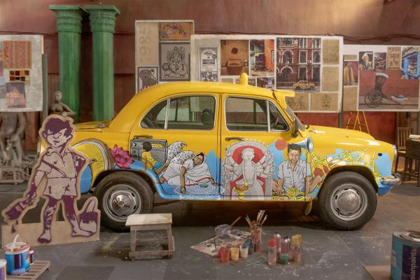 Choltey Choltey Chollish — Asian Paints A yellow vintage car, likely an Ambassador, is painted with intricate, colorful murals depicting various cultural scenes and figures, including a deity. The car is parked indoors, surrounded by art supplies like paint pots and brushes on the floor, and various artworks and posters are displayed on the wall in the background. A cardboard cutout of a boy with a paint bucket stands in the foreground.