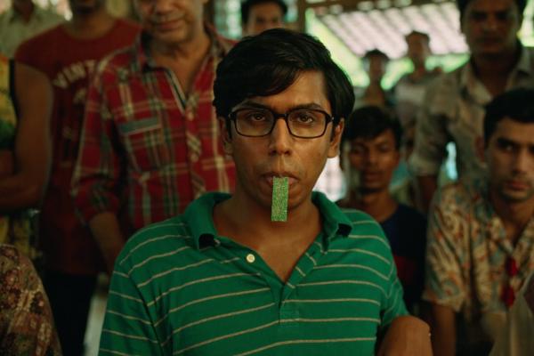 A young man with dark hair and glasses wears a green and white striped polo shirt, looking directly at the viewer with a green, rectangular object held between his lips. Several other people are blurred in the background.