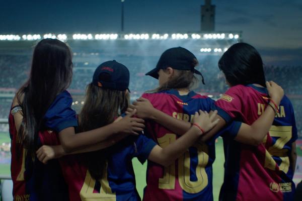 Four young girls, seen from behind, stand with their arms around each other's shoulders, watching a soccer game in a stadium at night. They are wearing red and blue soccer jerseys with numbers and names on the back. Two of the girls are wearing baseball caps. The stadium lights illuminate the field and the crowd in the background, creating a vibrant atmosphere.