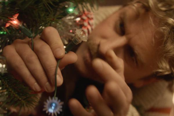 A close-up shot focuses on a person's hands holding a string of Christmas lights, attempting to fix a bulb. The person's face, with light-colored hair, is blurred in the background, appearing stressed or frustrated. The green branches of a Christmas tree are visible around the hands, along with other twinkling lights and tinsel.