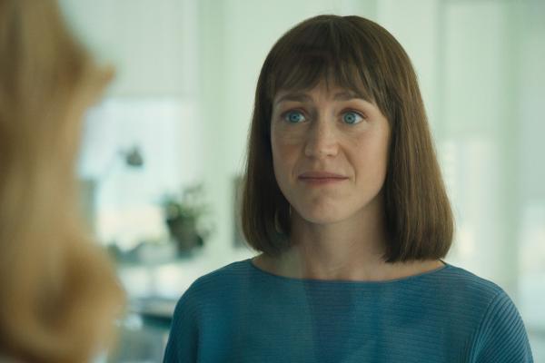 A woman with shoulder-length brown hair cut in a bob with bangs and striking blue eyes looks directly forward with a slightly concerned or serious expression. She is wearing a ribbed teal or blue top. Her gaze is focused, and her mouth is slightly open as if about to speak or having just spoken. The background is blurred, showing a bright, indoor environment, possibly an office, with light-colored walls and reflections.