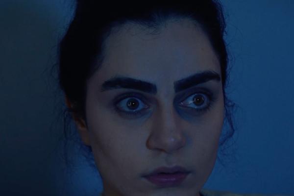 Halloween Essentials — Al-Futtaim IKEA A close-up of a woman's face with wide, startled eyes looking to the side. She has dark, prominent eyebrows and dark circles under her eyes, conveying a sense of fear or shock. The lighting is dim and casts a blue hue on her face.