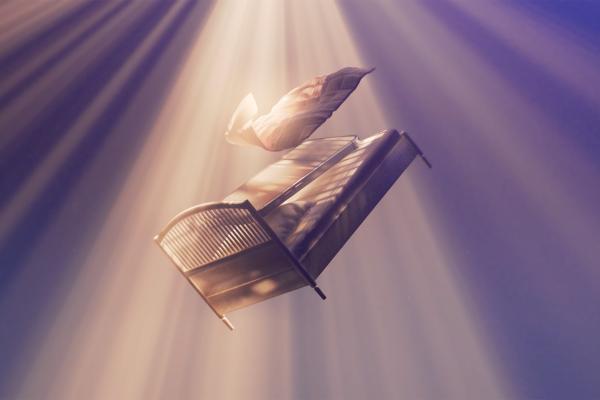 Phone Sleep Collection — IKEA A miniature white bed with a gray duvet and pillow sits on a white nightstand, next to a large black retro alarm clock. A black desk lamp hangs overhead, casting a dim light on the scene. A pair of eyeglasses rests on the nightstand near the bed.