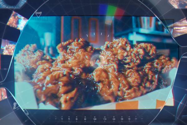 A close-up shot of several pieces of crispy, golden-brown fried chicken in a paper-lined basket. The image is viewed through a stylized, dark, circular frame with a central glowing gear-like icon and radiating white lines, resembling a futuristic lens or display. The background shows hints of a kitchen or restaurant setting.
