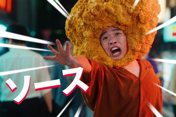 Sweet Tokyo — KFC A person wearing a large, textured, golden-brown costume on their head that resembles a piece of fried chicken. Their mouth is open in a wide expression, and one hand is outstretched. They are wearing an orange shirt. The background is a blurry, neon-lit street at night. Large Japanese text "ソース" (Sauce) is in the foreground, with white lines radiating from the person.