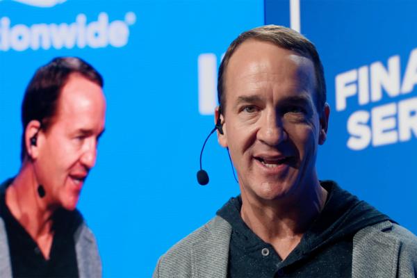 A close-up shot of Peyton Manning, a light-skinned man with short brown hair, smiling and speaking into a headset microphone. He is wearing a grey blazer over a dark hooded sweatshirt. His face is well-lit, and his eyes are looking slightly to the right. In the blurred background, a large blue screen shows the word "Nationwide" on the left and partially visible text "FINA SER" on the right, along with a reflection of Manning's image.