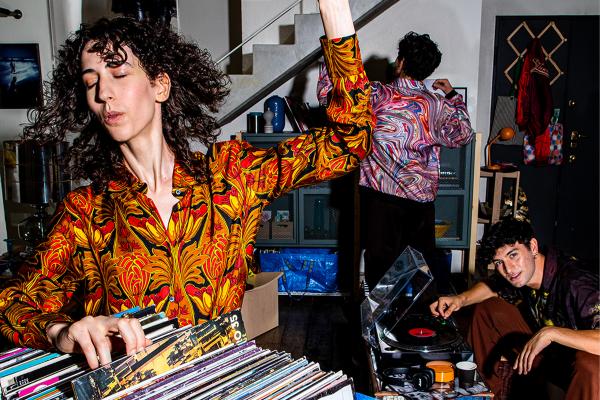 In an eclectic, warmly lit loft apartment, a woman with curly hair and a patterned shirt smiles as she flips through vinyl records, while friends dance and a DJ adjusts a turntable in the background.