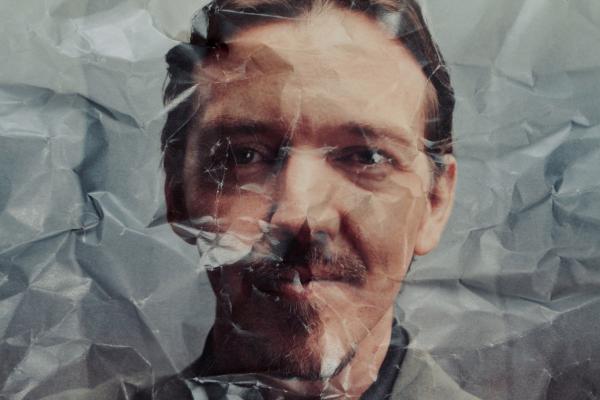 A portrait of a man with a mustache, looking directly at the viewer, obscured and distorted by a layer of severely crumpled, translucent, gray-white paper laid over the image. The wrinkles and folds of the paper are prominent across his face.