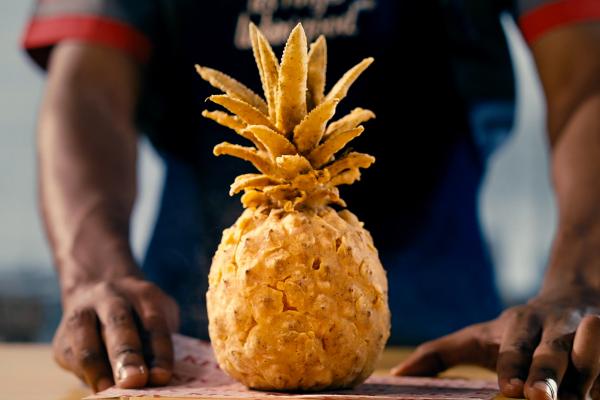 A pineapple covered in deep fried breading on a table in front of a person whose hands are on the table next to the pineapple