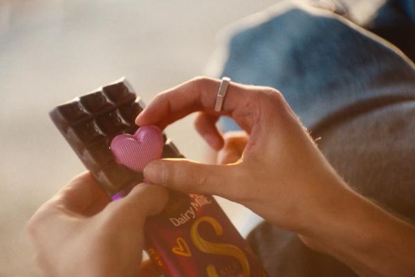 A close-up shot focusing on hands interacting with a chocolate bar. One hand holds an unwrapped dark chocolate bar, while the other hand, wearing a silver ring, is seen placing a small, ribbed, pink heart-shaped candy onto the chocolate. The purple wrapper of the Dairy Milk bar is partially visible in the bottom right corner.