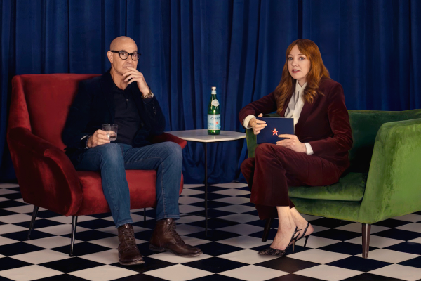 Stanley Tucci, in glasses, a dark blazer, and jeans, sits in a red armchair, looking towards Diane Morgan in a green armchair. Diane Morgan, with long reddish-brown hair and a burgundy velvet suit, holds a blue folder with a red star. A bottle of S.Pellegrino is on a small table between them. The background is a blue curtain, and the floor is black and white checkered.