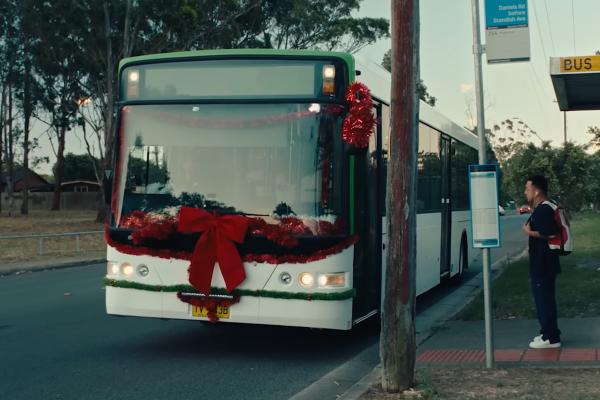 A white and green city bus, festively decorated with a large red bow and red tinsel around its front window, is stopped at a bus stop. A man with a backpack stands on the curb, looking towards the bus. The scene is set at dusk with trees and residential houses in the background.
