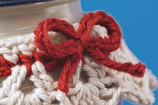 A close-up shot of a small red yarn bow tied on a cream and red crocheted cozy, which covers the top of a Heinz Mayonnaise jar. The background is a soft blue.