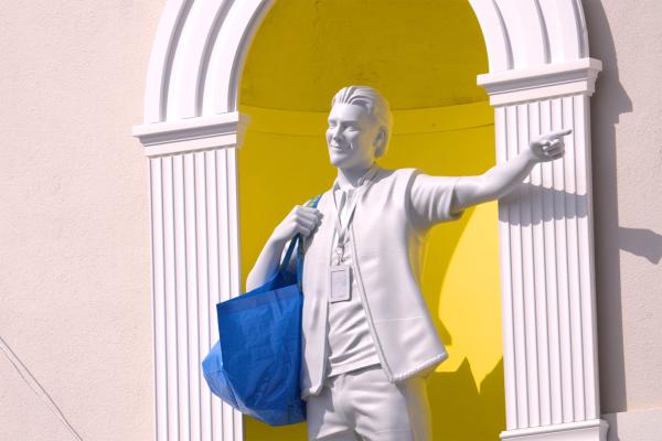 A close-up of a white, smiling statue of a man, wearing a casual t-shirt and vest, holding a blue IKEA Frakta shopping bag over his left shoulder. He is pointing forward with his right arm. The arched alcove behind the statue is painted a bright yellow, contrasting with the white architectural details.