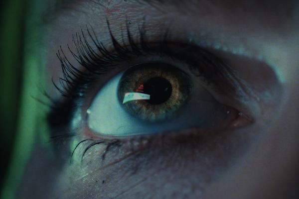 A close-up of a human eye with an intense, slightly upward gaze, bathed in a green and blue light. A small, bright reflection, possibly from a screen, is visible in the pupil.