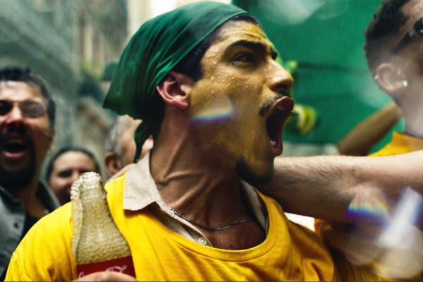 A close-up of a man with face paint and a green bandana, caught mid-shout with his mouth wide open. He is wearing a yellow shirt and has a glass Coca-Cola bottle at his shoulder. Raindrops and light refractions add to the vibrant, energetic atmosphere, with blurred faces of other people in the background.