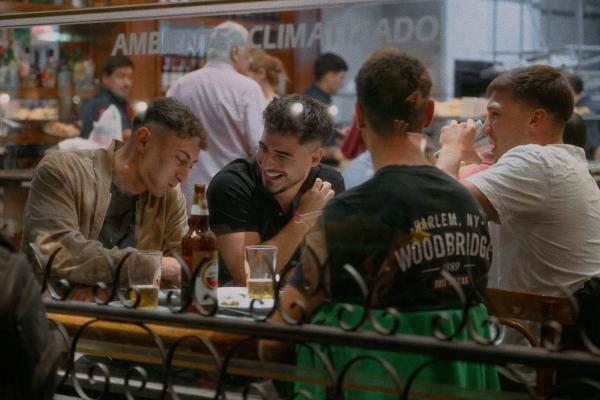 Four young men sitting at a table in a bustling restaurant or bar, seen through a glass partition. One man in a black shirt is laughing, while another in a beige jacket looks down. Two men are seen from behind, one wearing a black t-shirt that says "Harlem, NY Woodbridge." Bottles and glasses are on the table.