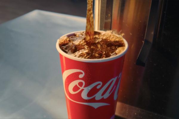 A red Coca-Cola paper cup filled with ice is being topped up with dark soda, causing bubbles and splashes. The dispenser is visible on the right, and the cup sits on a light-colored surface.