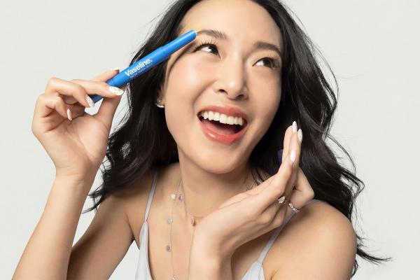 A joyful young Asian woman, with long dark hair, holding a blue Vaseline Brow Tamer product to her eyebrow, looking upwards and smiling broadly.