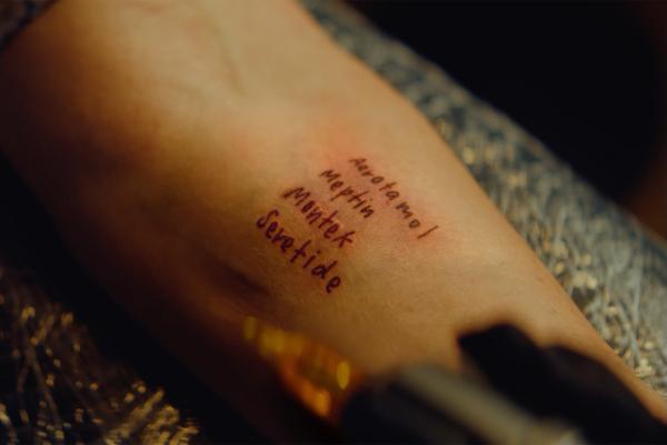 Close-up of a wrist or hand with handwritten tattoo-style text on the skin, lit warmly against a dark background.