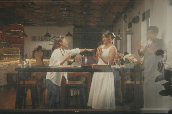 A view through a glass window showing two women inside a warm, modern cafe reacting dynamically to each other. One woman in a white dress holds her hands to her chest in delight while her friend in a white shirt and jeans points toward her excitedly, capturing a candid, playful moment of interaction in front of the cafe's counter area.