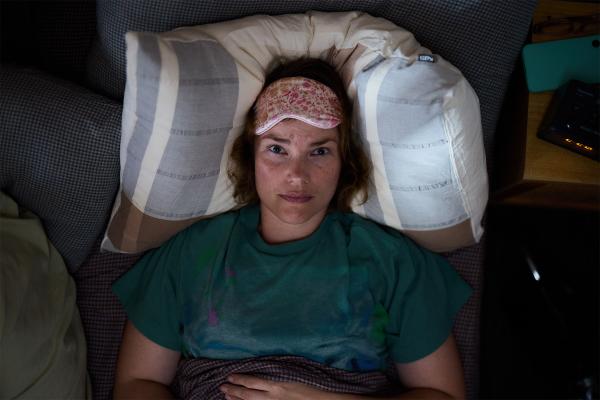 A top-down view of a woman lying awake in bed, resting her head on a striped pillow in a dimly lit room. She wears a floral sleep mask pushed up onto her forehead and stares directly at the camera with wide, weary eyes, conveying sleeplessness or insomnia.