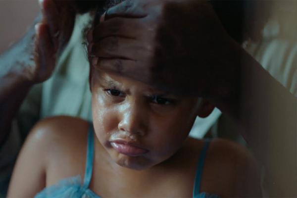 A close-up shot of a young child with a sulky expression as an adult's hands gently hold their head and smooth back their hair. The child wears a light blue tank top and looks down with pouting lips and furrowed brows.
