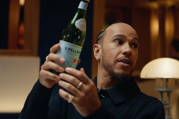 Lewis Hamilton holding up a green glass bottle of S.Pellegrino sparkling water with both hands while looking off-camera with a surprised, curious expression. He is wearing a dark jacket, diamond stud earrings, and several rings.