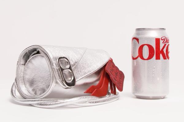 A cylindrical silver metallic handbag designed to resemble a soda can, featuring a silver pull-tab clasp and decorative red leather bag charms shaped like high heels and a "Diet Coke Break" tag. The purse rests on a white studio background next to a chilled, condensation-covered Diet Coke can for scale.