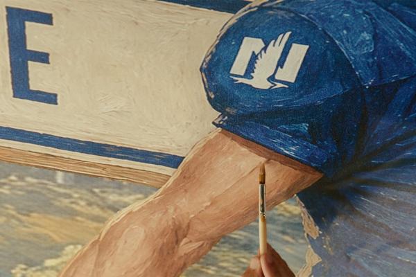 Close-up of a paintbrush adding detail to a mural of a football player’s arm beneath a blue jersey sleeve bearing the Nationwide eagle logo.