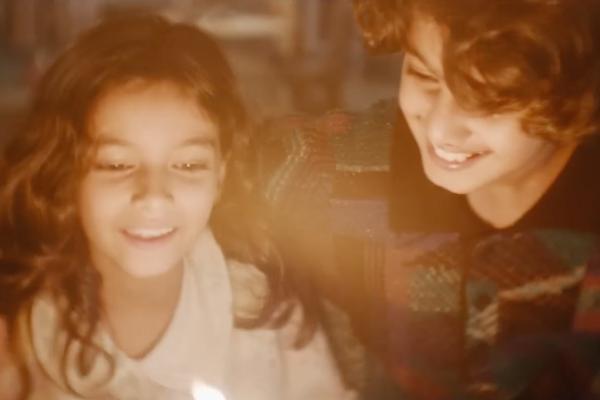 Close-up of a young girl with long wavy brown hair and an older boy with curly hair, both smiling warmly as their faces are illuminated by a soft, golden light from a candle flame below.