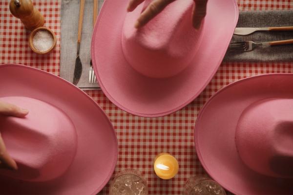 Top-down view of a dinner table set with a red-and-white gingham tablecloth, cutlery, and a small candle, where three upside-down pink cowboy hats serve as dish covers as hands reach in to reveal what is beneath.
