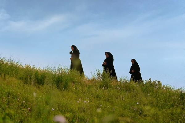 Wide shot of three women wearing traditional black long dresses and black headscarves walking in single file along the crest of a lush green hill with wildflowers under a clear blue sky.
