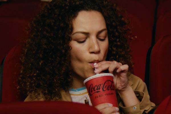 Close-up of a young woman with curly hair seated in red cinema chairs, sipping from a red Coca-Cola Zero Sugar cup.