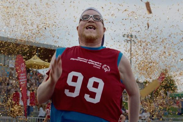 Close-up of a runner wearing a red Special Olympics Canada jersey racing across the finish line as sparkling gold confetti showers down against an outdoor stadium backdrop.
