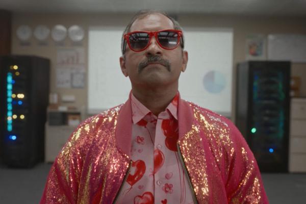 middle aged man with a mustache wearing red sunglasses, a pink glittering jacket and a pink button up shirt with hearts on it 