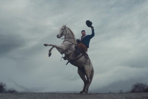 A man with a mustache, wearing a blue jacket and brown chaps, rides a white horse that is rearing up on its hind legs. The man is raising his hat in the air, appearing celebratory, against a backdrop of a cloudy, grey sky and a barren landscape.