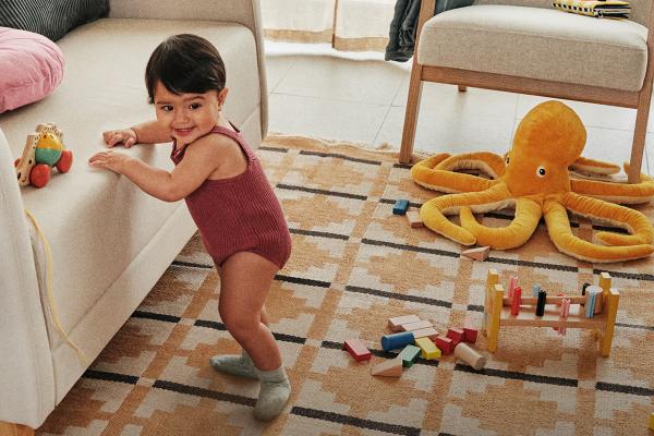 A baby stands with the help of a couch. On the floor are kids toys -- wooden blocks and a stuffed animal octopus