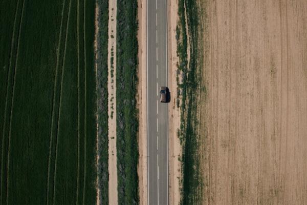 Bird's eye view of a single car on a road, with brown dirt on one side of the road and green grass/trees on the other  