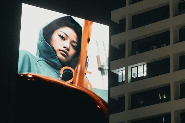 A billboard outside at night. On the billboard, coffee flows down into a mug. Behind the liquid, a woman wearing a jacket with a hood.