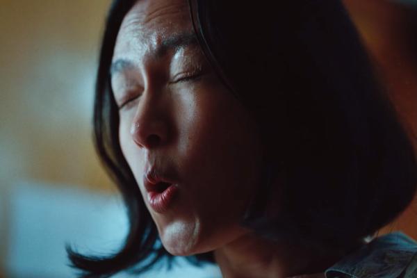 A close-up shot of a person's face, with their eyes closed and mouth slightly open as if exhaling or making a sound. Their skin appears slightly sweaty or dewy, and their dark hair frames their face. The background is blurred with hints of light and shadow.