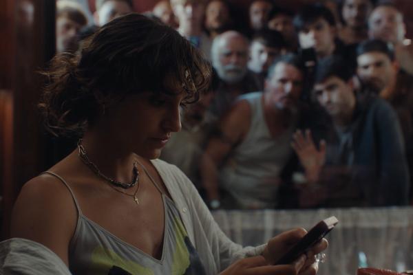 A young woman with dark, curly hair looks down at a phone in her hands, wearing a light-colored top and a cardigan, with a cross necklace visible. Behind her, a blurred crowd of people, mostly men, are visible, some looking towards her.